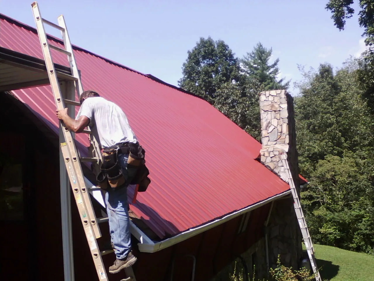 Roofer on a ladder beside a red metal roof and stone chimney — Soffit & Fascia Repair by Roofing Brothers in Diamond